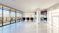 This expansive single-floor amenity space offers a thoughtfully designed environment that blends comfort, functionality, and contemporary style. The open layout is anchored by polished white floors that reflect abundant natural light streaming in through large, floor-to-ceiling windows, which frame sweeping panoramic city views from what appears to be an elevated position within the building.

The space is organized into several distinct zones. A generous open lounge area features stylish sofas, accent chairs, and coffee tables arranged atop patterned rugs, complemented by a striking marble-surround fireplace and warm wood-paneled ceiling with exposed beams. Adjacent to this is a fully equipped kitchen bar with counter seating, pendant lighting, and built-in shelving — ideal for social gatherings.

A quieter area provides privacy-focused seating with high-back acoustic chair pods positioned along a textured feature wall, offering an intimate setting within the broader open space. A separate lounge nook includes a wall-mounted television, a sectional sofa, and curated artwork, creating a more relaxed retreat.

Entry points feature warm natural stone wall cladding, glass double doors, and direct access to what appears to be an outdoor terrace. Throughout, the design balances natural wood tones, white walls, and modern furnishings to create a welcoming, upscale communal atmosphere.
