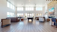 This expansive single-floor community space showcases a contemporary design highlighted by soaring double-height ceilings and an abundance of natural light streaming through large clerestory and ground-level windows along multiple walls. The flooring throughout is a light-toned wood-look plank, lending warmth and continuity across the open layout.

The main lounge area is generously proportioned, featuring distinct seating zones with a mix of leather sofas, accent chairs, and a central coffee table arrangement set atop patterned area rugs. A billiards table anchors the game area at the heart of the space. Adjacent to the lounge, a built-in counter with bar-height seating in bold blue and orange tones is positioned beside a dramatic floor-to-ceiling stacked stone fireplace, which serves as a focal architectural centerpiece. Decorative wood beam detailing overhead adds a layer of texture and visual interest.

A dedicated work nook lines one windowed wall, outfitted with bar-height desks and chairs, providing a functional workspace with outdoor views. A separate private meeting or conference room, accessible through French doors with frosted glass sidelights, offers a more intimate setting with patterned carpet, dark cabinetry, and curated wall art.

The entry vestibule features glazed double doors flanked by vertical wall sconces and potted greenery, creating a welcoming arrival experience. Cozy seating alcoves throughout complete this well-appointed communal environment.