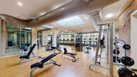 This expansive fitness center is organized across a single floor into several distinct functional zones, each designed to support a comprehensive workout experience. The main strength training area features exposed brick walls, warm hardwood-style flooring, and prominent exposed ductwork overhead, lending an industrial-modern aesthetic. This zone is equipped with cable machines, power racks, adjustable benches, a full dumbbell rack, and free weights, with large mirrors enhancing the sense of space.

A dedicated cardio area lines floor-to-ceiling windows, flooding the space with natural light and offering outdoor views. This section houses a row of treadmills, ellipticals, stationary bikes—including recumbent and upright models—and rowing machines. The bright, airy atmosphere makes this zone particularly inviting.

A separate functional training room features a bold accent wall, wall-mounted television, floor-to-ceiling mirrors, and an open layout accommodating balance equipment, kettlebells, resistance bands, a boxing bag, medicine balls, and exercise mats—ideal for group or individual training sessions.

Throughout, recessed and panel lighting ensures consistent illumination, while the cohesive hardwood flooring unifies the various zones into a well-appointed, professional-grade fitness facility.