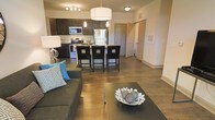 This single-floor residence offers a thoughtfully designed open-concept layout that seamlessly connects the living, dining, and kitchen areas into one cohesive and functional space. Rich hardwood-style flooring flows throughout, anchoring the modern aesthetic and creating a warm, inviting ambiance.

The living area is comfortably appointed, featuring a plush sofa accented with decorative throw pillows and a sleek dark coffee table. A statement pendant lamp adds a refined touch to the space. The kitchen is a standout feature, showcasing dark espresso cabinetry, a full suite of stainless steel appliances including a refrigerator, stove, and microwave, and a stylish tile backsplash. A kitchen island with bar-height seating for multiple guests makes the space ideal for both casual dining and entertaining.

Overhead, a combination of recessed track lighting, pendant fixtures, and a bold drum pendant light above the dining area ensures the space is well-lit and visually dynamic. Doors visible in the background hint at additional rooms beyond the main living area, including what appears to be a utility or storage space. The overall design blends contemporary style with practical functionality, making this a polished and comfortable single-floor residence.