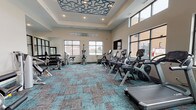Located on Bushwood Drive in Elgin, IL, this well-appointed fitness center offers a comprehensive workout environment designed with both function and aesthetics in mind. The single-floor space is defined by soaring ceilings accented with an intricate teal decorative ceiling medallion, recessed lighting, and a raised tray ceiling detail that adds architectural character to the room.

Natural light floods the interior through large, dark-framed windows positioned along multiple walls, providing open views of the surrounding community. The floor is finished with a stylish teal and gray patterned carpet tile that ties together the modern color palette throughout the space.

The cardio zone features multiple treadmills, elliptical trainers, and stationary cycles, including Cybex-branded equipment, complemented by a wall-mounted flat-screen television for entertainment during workouts. The strength training area includes a full dumbbell rack with a variety of weights, an adjustable weight bench, and several Hoist-branded selectorized strength machines targeting key muscle groups. Floor-to-ceiling glass partition walls with frosted accents separate areas within the space, adding visual openness while defining distinct workout zones. Full-length mirrors enhance the sense of space in the free weights section. A small storage shelving unit provides convenient organization for personal items or fitness accessories.