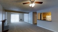 This single-floor unit features an open-concept living area finished with wall-to-wall carpeting in a neutral tone, creating a comfortable and cohesive feel throughout the space. A ceiling fan with an integrated light fixture is centrally mounted on the textured popcorn ceiling, providing both airflow and ambient lighting. A large window fitted with vertical blinds allows natural light to fill the room while offering privacy control. A wall-mounted HVAC unit sits beneath the window, providing supplemental climate control. The living area flows seamlessly toward a pass-through counter that connects to the kitchen, offering a practical and social link between the two spaces. The kitchen, partially visible from the living area, features warm wood-toned cabinetry and a dark countertop surface. The layout is straightforward and functional, with clean white baseboards framing the neutral gray walls. The overall ambiance is understated and move-in ready, offering a blank canvas well-suited for a variety of furnishing styles.