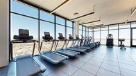 Located in Phoenix, AZ, this high-rise fitness center offers a generously sized, modern workout facility designed to meet a wide range of fitness needs. The space is defined by dramatic floor-to-ceiling glass curtain walls that flood the interior with natural light and provide sweeping panoramic views of the Phoenix skyline and surrounding desert landscape, including distant mountain ranges.

The main fitness floor is finished with large-format dark tile and houses an extensive array of cardio equipment, including multiple treadmills, elliptical trainers, stair climbers, and stationary bikes. A dedicated strength training area features a full rack of dumbbells, weight benches, cable machines, and functional training equipment, complemented by wall-mounted mirrors and mounted flat-screen televisions.

A separate studio room offers a distinct environment for yoga, stretching, or group fitness classes. This area features light hardwood flooring, elegant circular ceiling light fixtures, and its own floor-to-ceiling glass doors that open to views of the city, with floor-length drapes providing privacy when needed.

Throughout the facility, linear pendant lighting and recessed fixtures create a bright, clean aesthetic. The overall design is contemporary and functional, balancing high-end finishes with practical athletic utility in an elevated urban setting.