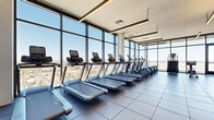 Located in Phoenix, AZ, this high-rise fitness center offers a generously sized, modern workout facility designed to meet a wide range of fitness needs. The space is defined by dramatic floor-to-ceiling glass curtain walls that flood the interior with natural light and provide sweeping panoramic views of the Phoenix skyline and surrounding desert landscape, including distant mountain ranges.

The main fitness floor is finished with large-format dark tile and houses an extensive array of cardio equipment, including multiple treadmills, elliptical trainers, stair climbers, and stationary bikes. A dedicated strength training area features a full rack of dumbbells, weight benches, cable machines, and functional training equipment, complemented by wall-mounted mirrors and mounted flat-screen televisions.

A separate studio room offers a distinct environment for yoga, stretching, or group fitness classes. This area features light hardwood flooring, elegant circular ceiling light fixtures, and its own floor-to-ceiling glass doors that open to views of the city, with floor-length drapes providing privacy when needed.

Throughout the facility, linear pendant lighting and recessed fixtures create a bright, clean aesthetic. The overall design is contemporary and functional, balancing high-end finishes with practical athletic utility in an elevated urban setting.