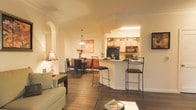 This single-floor residence showcases an open-concept layout that effortlessly connects the living room, dining area, and kitchen into one cohesive and inviting space. Rich hardwood flooring runs throughout, anchoring the warm, neutral color palette that gives the interior a welcoming ambiance.

The living room offers a comfortable, well-lit setting with soft natural tones and easy access to the rest of the space. A graceful arched entryway adds an architectural detail that enhances the flow between rooms.

The dining area features a round table with dark upholstered chairs, set beneath a pendant light fixture, and is complemented by a bold red accent wall that creates a vibrant focal point. Decorative wall art and a floral centerpiece add warmth and personality to the space.

The kitchen is designed for both function and style, featuring light wood cabinetry, a black refrigerator, a built-in microwave, and a gas range. A curved breakfast bar with wrought iron bar stools bridges the kitchen and living areas, offering casual seating and enhancing the open, social feel of the layout. Crown molding throughout the space adds a refined finishing touch, elevating the overall aesthetic.