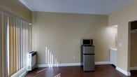 This single-floor space presents a clean, minimalist layout with a focus on functional simplicity. The room features warm beige walls that create a neutral, inviting atmosphere, complemented by rich dark hardwood flooring that runs throughout the space. White baseboards provide a crisp contrast to both the walls and flooring, contributing to a tidy, well-defined interior.

Natural light enters through a window on the left side of the room, casting distinct light patterns across the floor and adding brightness to the otherwise understated space. A wall-mounted heating unit is positioned near the window, offering a practical climate control solution.

The compact kitchen area is equipped with a stainless steel mini refrigerator topped with a microwave and an additional small appliance, neatly arranged against the far wall. This setup provides essential kitchen functionality within a modest footprint.

The space is currently unfurnished, allowing the architectural elements — including the smooth white ceiling and clean wall surfaces — to stand out. The overall ambiance is simple and utilitarian, suitable for a variety of uses.