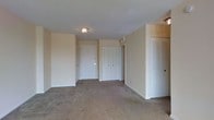 This single-floor space presents a straightforward, functional layout finished in a neutral color palette throughout. The main living area is covered in wall-to-wall carpeting in a warm beige tone, providing a soft underfoot feel that extends across the open floor plan. Walls are painted in a light, understated hue that contributes to a clean and airy atmosphere.

A flush-mount ceiling light fixture provides overhead illumination in the central area, complementing the ambient light present in the space. The entryway features a solid white door, offering a defined point of entry into the unit.

Storage is a notable feature of this space, with multiple closet options visible throughout. A set of bifold doors conceals what appears to be a generously sized closet, while louvered closet doors on the opposing side provide additional dedicated storage. A secondary interior door suggests access to an adjoining room or hallway, adding to the overall functionality of the layout.

The white trim work and door frames provide clean contrast against the neutral walls, maintaining a cohesive and tidy aesthetic throughout. The space offers a versatile, move-in-ready foundation suitable for a variety of furnishing arrangements.