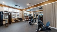 Located at 20 Franklin in Manalapan, NJ, this fitness center is a thoughtfully designed exercise space offering a comprehensive range of workout equipment and a polished, modern aesthetic. The room features durable rubber flooring throughout, providing a safe and practical surface for all types of training activities.

The space is outfitted with a full suite of professional-grade equipment, including multiple treadmills, an elliptical, a rowing machine, a multi-function cable/strength machine, and a dedicated free weights station with a full rack of dumbbells. Medicine balls and stability balls round out the functional training options.

Architecturally, the room is distinguished by crown molding, a warm gold accent band along the upper walls, and vertical wood slat wall panels that add a contemporary design element. A full-length mirror along one wall visually expands the space and supports proper form during workouts. Recessed lighting and ceiling fans with sleek black blades ensure the room remains well-lit and comfortably ventilated. Large windows on multiple walls flood the space with natural light and offer views of the surrounding area.

A wall-mounted TV is positioned for convenient viewing during cardio sessions. The space is accessed through elegant black-framed French doors, adding a refined touch to the overall design. This fitness center is a fully functional and attractively appointed facility suited for a wide range of workout routines.