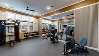 Located at 20 Franklin in Manalapan, NJ, this fitness center is a thoughtfully designed exercise space offering a comprehensive range of workout equipment and a polished, modern aesthetic. The room features durable rubber flooring throughout, providing a safe and practical surface for all types of training activities.

The space is outfitted with a full suite of professional-grade equipment, including multiple treadmills, an elliptical, a rowing machine, a multi-function cable/strength machine, and a dedicated free weights station with a full rack of dumbbells. Medicine balls and stability balls round out the functional training options.

Architecturally, the room is distinguished by crown molding, a warm gold accent band along the upper walls, and vertical wood slat wall panels that add a contemporary design element. A full-length mirror along one wall visually expands the space and supports proper form during workouts. Recessed lighting and ceiling fans with sleek black blades ensure the room remains well-lit and comfortably ventilated. Large windows on multiple walls flood the space with natural light and offer views of the surrounding area.

A wall-mounted TV is positioned for convenient viewing during cardio sessions. The space is accessed through elegant black-framed French doors, adding a refined touch to the overall design. This fitness center is a fully functional and attractively appointed facility suited for a wide range of workout routines.