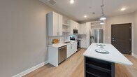 Located at 1006 E Broad St in Savannah, Georgia, this single-floor unit offers a thoughtfully designed open layout with contemporary finishes throughout. The living area features warm wood-look luxury vinyl plank flooring and a ceiling fan, creating a bright and comfortable space that flows seamlessly into the kitchen and adjoining rooms.

The kitchen is a standout feature, outfitted with crisp white shaker-style cabinetry, gleaming quartz countertops, and a striking hexagonal tile backsplash. Stainless steel appliances—including a range, over-the-range microwave, dishwasher, and refrigerator—complete the space, while pendant lighting and open shelving add a modern, functional touch. A dark-finished island provides additional prep and dining space.

The bedroom is carpeted for comfort and receives ample natural light through a pair of large windows dressed with horizontal blinds, offering views of the surrounding greenery. A ceiling fan adds to the room's practicality.

The bathroom presents a clean, spa-like aesthetic with wood-look tile flooring, a generous vanity with white cabinetry and an oversized mirror, and well-placed lighting. A dedicated laundry area houses a full-size top-load washer and dryer with overhead shelving, providing excellent in-unit convenience. The overall palette of soft grays and whites throughout creates a cohesive, modern ambiance.