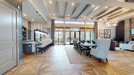 Located at 8050 Lapis Ln NW in Concord, NC, this single-floor clubhouse facility showcases an upscale, open-concept design that seamlessly blends modern sophistication with warm, rustic elements. The grand entryway sets the tone with striking geometric parquet hardwood flooring, soaring ceilings, and a wide corridor that draws the eye toward floor-to-ceiling windows and glass doors opening to an outdoor patio area. Throughout the space, exposed reclaimed wood ceiling beams add character and contrast beautifully against the crisp white coffered ceiling details and recessed lighting.

The main gathering area features a plush lounge with a statement stone fireplace, a wall-mounted TV, and comfortable seating arranged for both conversation and relaxation. Adjacent to this is a spacious dining area centered around a large dark wood table with upholstered seating, perfectly positioned beneath pendant lighting and framed by expansive glass doors that invite natural light and outdoor views.

The kitchen and bar area is a standout feature, offering an extended granite-topped island with patterned upholstered barstools, sleek gray cabinetry, floating wood shelves, and an arched stone accent wall with a mounted display screen. A curated gallery wall and decorative framed artwork complete the refined, welcoming atmosphere of this impressive community space.