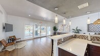 Located in Phoenix, Arizona, this well-appointed single-floor community space offers an open-concept layout that seamlessly connects the kitchen, dining room, and living room. Warm wood-look flooring flows throughout, complemented by crisp white walls and recessed lighting that keep the interior bright and inviting.

The kitchen features a spacious peninsula with a white quartz countertop accented by a mosaic tile backsplash and dark cabinetry. Pendant lights hang above the bar-height seating, adding a modern touch. Stainless steel appliances and ample counter space make it a highly functional area.

The living room is airy and welcoming, anchored by a tufted sectional sofa and a round coffee table. Large-scale abstract artwork adorns the walls, adding a contemporary flair. A wall-mounted flat-screen TV and a pair of leather accent chairs create a secondary lounge area near the glass French doors, which open directly to an outdoor pool and patio — flooding the space with natural light and offering seamless indoor-outdoor connectivity.

The dining area features a round table beneath a woven pendant light, accompanied by a wall-mounted oversized Scrabble board that adds a playful, social element. A dedicated counter station near the kitchen provides additional workspace. The overall ambiance is modern, clean, and community-focused, ideal for gathering and relaxation.