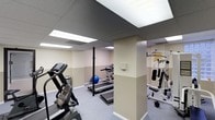 This fitness center offers a practical and well-organized space designed to support a full range of workout routines. The room features a drop ceiling with recessed panel lighting that provides consistent, bright illumination throughout the area. The walls are finished in a two-tone neutral palette — a lighter upper half paired with a warm beige lower section — lending the space a clean and functional aesthetic. Durable low-pile carpet covers the floor, offering comfort underfoot during exercise.

The cardio zone is well-appointed, featuring a treadmill and a stair climber with digital displays, positioned for easy use. A structural column serves as a natural divider within the open layout without interrupting the flow of the space. Toward the back, an additional treadmill and a stability ball suggest dedicated space for stretching and core training. On the opposite side, a multi-station cable and weight machine provides options for full-body strength training, accompanied by free weights nearby. An informational bulletin board is mounted on the wall, offering guidance or facility notices for users.

Overall, the space is thoughtfully arranged to maximize functionality, providing a comfortable and accessible environment for both cardio and strength-focused workouts.