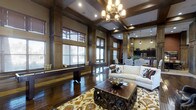This sophisticated single-floor space is defined by its rich architectural character and refined design details. Dark hardwood flooring extends throughout, complemented by floor-to-ceiling wood-paneled walls and ornate coffered ceilings that lend a sense of grandeur and depth to the interior. A statement chandelier anchors the main living area, where a plush white sofa, an intricately carved wooden coffee table, and a vibrant patterned area rug create a warm and inviting focal point. Large windows dressed with wooden blinds line the walls, allowing natural light to filter in while maintaining a sense of privacy. Adjacent to the living area, a game room features a shuffleboard table set against a built-in bench, adding a recreational element to the space. The open-concept layout flows naturally into a dining area and a well-appointed kitchen, which is outfitted with dark cabinetry, pendant lighting, and modern appliances. Recessed lighting and decorative pendants throughout ensure a warm, layered ambiance. The cohesive use of dark wood tones, thoughtful furnishings, and architectural detailing gives this space a distinguished, club-like atmosphere that is both functional and visually impressive.