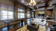This sophisticated single-floor space is defined by its rich architectural character and refined design details. Dark hardwood flooring extends throughout, complemented by floor-to-ceiling wood-paneled walls and ornate coffered ceilings that lend a sense of grandeur and depth to the interior. A statement chandelier anchors the main living area, where a plush white sofa, an intricately carved wooden coffee table, and a vibrant patterned area rug create a warm and inviting focal point. Large windows dressed with wooden blinds line the walls, allowing natural light to filter in while maintaining a sense of privacy. Adjacent to the living area, a game room features a shuffleboard table set against a built-in bench, adding a recreational element to the space. The open-concept layout flows naturally into a dining area and a well-appointed kitchen, which is outfitted with dark cabinetry, pendant lighting, and modern appliances. Recessed lighting and decorative pendants throughout ensure a warm, layered ambiance. The cohesive use of dark wood tones, thoughtful furnishings, and architectural detailing gives this space a distinguished, club-like atmosphere that is both functional and visually impressive.