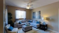 This single-floor space offers a thoughtfully designed living area that blends comfort and contemporary style. The living room serves as the centerpiece, featuring a large L-shaped sectional sofa in soft grey upholstery, accented with teal and blue decorative pillows that add a vibrant pop of color. A matching upholstered ottoman serves as both a coffee table and additional seating, anchoring the space over a plush area rug. The room is bathed in warm, ambient light from a classic ceiling fan with an integrated light fixture, as well as a stylish table lamp that casts a soft glow in the corner. A window dressed with sheer curtains allows natural light to filter in, offering a view of greenery outside. The walls are painted in a warm taupe tone, complemented by a striking multi-panel abstract artwork in blue and grey hues, adding an artistic focal point to the room. A dark wood side table with a lower shelf provides functional storage and display space. The overall ambiance is relaxed yet polished, making it a welcoming environment suited for both everyday living and entertaining.