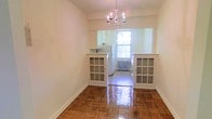 This single-floor space showcases a warm, classic aesthetic defined by richly polished parquet hardwood flooring that extends throughout the main living and dining areas. An elegant chandelier serves as a focal point overhead, adding a timeless charm to the dining space. The room is framed by crisp white walls that lend a clean, airy feel to the interior.

A distinctive architectural detail is the built-in pass-through opening that connects the dining area to the kitchen, flanked by two white-framed glass-door display cabinets. These cabinets add both storage and visual character, creating a charming transitional zone between the spaces. The kitchen beyond features white cabinetry and appliances, maintaining a cohesive, light color palette throughout. A window in the kitchen allows natural light to pour into the space, brightening both rooms and offering a glimpse of greenery outside. A classic radiator beneath the window indicates a traditional heating system. The overall ambiance is cozy yet refined, blending vintage architectural elements with practical functionality in a well-maintained, light-filled layout.