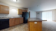 This single-floor unit offers a functional and well-organized layout with a cohesive design throughout. The kitchen serves as a central highlight, featuring warm honey-toned wood cabinetry, granite-look countertops, and a full suite of black appliances including a refrigerator, electric range, built-in microwave, and dishwasher. Stone-look tile flooring anchors the kitchen, which also includes a center island with additional counter space and a convenient electrical outlet. The kitchen opens directly into the living area, creating an easy flow between spaces.

The living area is generously sized, finished with plush neutral carpet and baseboard heating. Natural light enters through a window dressed with vertical blinds, and a wall-mounted AC unit provides climate control. Bedrooms continue the carpeted flooring and feature baseboard heaters and windows with blinds, offering comfortable, private retreats.

The bathrooms are finished with the same stone-look tile flooring and include warm wood vanities topped with granite-look countertops. One bathroom includes an adjacent closet with wire shelving for added storage. A dedicated laundry area houses a full-size washer and dryer, completing this practical and move-in-ready unit.