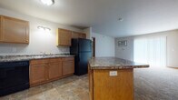 This single-floor unit offers a functional and well-organized layout with a cohesive design throughout. The kitchen serves as a central highlight, featuring warm honey-toned wood cabinetry, granite-look countertops, and a full suite of black appliances including a refrigerator, electric range, built-in microwave, and dishwasher. Stone-look tile flooring anchors the kitchen, which also includes a center island with additional counter space and a convenient electrical outlet. The kitchen opens directly into the living area, creating an easy flow between spaces.

The living area is generously sized, finished with plush neutral carpet and baseboard heating. Natural light enters through a window dressed with vertical blinds, and a wall-mounted AC unit provides climate control. Bedrooms continue the carpeted flooring and feature baseboard heaters and windows with blinds, offering comfortable, private retreats.

The bathrooms are finished with the same stone-look tile flooring and include warm wood vanities topped with granite-look countertops. One bathroom includes an adjacent closet with wire shelving for added storage. A dedicated laundry area houses a full-size washer and dryer, completing this practical and move-in-ready unit.
