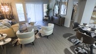 This thoughtfully designed single-floor space showcases a seamlessly flowing open-concept layout that combines comfort with contemporary style. The living area is anchored by rich, grey-toned hardwood-style flooring that extends throughout, providing a cohesive and elegant foundation for the entire space.

The main living area is generously proportioned, featuring a curated seating arrangement with sleek, cream-colored accent chairs and a patterned area rug that adds visual depth and warmth. Large windows dressed with vertical blinds allow abundant natural light to fill the room, creating a bright and airy ambiance throughout the day.

A stylish wooden credenza serves as a focal point along one wall, topped with decorative accents and framed by a striking pair of circular wall art pieces, lending the space a modern, curated aesthetic. Adjacent to the main seating area, a dining space accommodates a patterned table surrounded by chairs, ideal for casual meals or entertaining.

Lush indoor greenery, including a tall tropical plant near the window, introduces a refreshing natural element that complements the neutral color palette of greys, whites, and warm wood tones. The overall ambiance is modern yet inviting, blending clean architectural lines with carefully chosen furnishings to create a space that is both functional and visually refined.