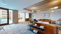 This thoughtfully designed single-floor amenity space offers a seamless blend of modern aesthetics and functional comfort across three distinct zones. The main lounge area is anchored by a striking wood-paneled tray ceiling, plush patterned carpeting, and rich cognac leather sofas complemented by navy accent chairs. Built-in shelving and curated artwork add warmth and character, while a bold textured leather tile feature wall in the entry corridor sets a sophisticated tone from the moment you arrive. The media room continues the warm wood ceiling detail and introduces a tiled accent wall as a backdrop for a large flat-screen television, flanked by a pair of leather sofas and round marble-topped coffee tables — ideal for communal gatherings. The dining and social area transitions to polished terrazzo flooring and is bathed in abundant natural light through floor-to-ceiling glass doors that open to views of an outdoor pool terrace. This zone features multiple seating configurations, including a round wooden dining table and a long communal table with leather chairs, as well as counter-height bar seating. A vaulted ribbed ceiling with sculptural detailing adds architectural interest overhead. A private dining room with an arched coffered ceiling, large-format artwork, and an eight-seat table rounds out the space with an intimate, refined atmosphere.