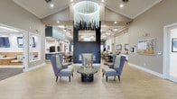 This single-floor space makes an immediate impression with its grand, open-concept lobby, anchored by a dramatic vaulted ceiling and a stunning cascading chandelier that serves as the room's centerpiece. The soaring ceiling height lends a sense of grandeur and airiness throughout the main gathering area.

The lounge area is thoughtfully arranged with plush, high-backed upholstered chairs in muted blue and grey tones surrounding a sculptural round coffee table, creating an inviting space for conversation and relaxation. A sleek, modern fireplace set against a bold dark accent wall adds warmth and a focal point to the seating arrangement, framed above by a contemporary circular light fixture.

Light wood-toned flooring runs throughout, complementing the neutral palette of warm beige and deep teal accent walls. Recessed lighting and ceiling fans are distributed evenly across the space, ensuring both comfort and well-lit functionality.

Adjacent to the lounge, a dining area is visible, furnished with neatly arranged tables and chairs beneath additional ceiling fans, suggesting a communal space well-suited for group gatherings. A reception or service counter is visible toward the back, indicating a staffed, hospitality-oriented environment. Framed artwork adorns the walls, adding character and a curated aesthetic throughout this welcoming, multifunctional facility.