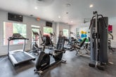 This fitness center is thoughtfully designed to support a full range of workout activities across a single level. The space is divided into distinct functional zones, beginning with a strength training area outfitted with a multi-station cable machine, adjustable weight benches, and a tiered dumbbell rack stocked with a complete set of free weights. Floor-to-ceiling mirrors line one wall, amplifying the sense of space and allowing users to monitor their form during exercise. The cardio zone features a treadmill, elliptical trainer, stair climber, and a rowing machine, positioned to take advantage of natural light streaming through large windows that offer views of an outdoor courtyard area. A wall-mounted flat-screen television adds convenience for cardio sessions. An adjoining studio space provides a clean, open floor plan with a mounted ballet barre and mirrored wall, making it well-suited for yoga, stretching, pilates, or group fitness classes. Colorful exercise balls and foam rollers are visible within this secondary room. Throughout the facility, durable dark rubberized flooring provides comfort and safety underfoot. The clean white walls, recessed lighting, and ceiling fans contribute to a bright, modern, and well-ventilated atmosphere conducive to focused training.