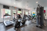 This fitness center is thoughtfully designed to support a full range of workout activities across a single level. The space is divided into distinct functional zones, beginning with a strength training area outfitted with a multi-station cable machine, adjustable weight benches, and a tiered dumbbell rack stocked with a complete set of free weights. Floor-to-ceiling mirrors line one wall, amplifying the sense of space and allowing users to monitor their form during exercise. The cardio zone features a treadmill, elliptical trainer, stair climber, and a rowing machine, positioned to take advantage of natural light streaming through large windows that offer views of an outdoor courtyard area. A wall-mounted flat-screen television adds convenience for cardio sessions. An adjoining studio space provides a clean, open floor plan with a mounted ballet barre and mirrored wall, making it well-suited for yoga, stretching, pilates, or group fitness classes. Colorful exercise balls and foam rollers are visible within this secondary room. Throughout the facility, durable dark rubberized flooring provides comfort and safety underfoot. The clean white walls, recessed lighting, and ceiling fans contribute to a bright, modern, and well-ventilated atmosphere conducive to focused training.