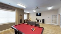 Located at 150 Horizon Trail in Garner, NC, this single-floor space is designed to blend entertainment and relaxation seamlessly. The open-concept layout centers around a full-size pool table with a bold red felt surface, complemented by a wall-mounted cue rack — making it an ideal gathering spot for recreational activities. Flush against one wall, a wall-mounted flat-screen TV sits above a rustic media console, anchoring a comfortable lounge area furnished with a sectional sofa and additional seating. The space benefits from abundant artificial lighting provided by multiple flush-mount ceiling light panels, as well as a ceiling fan that contributes to air circulation and comfort. Natural light filters in through multiple windows dressed with neutral-toned curtains, adding warmth to the room's earthy color palette of taupe and beige tones. Light wood-look flooring runs throughout, enhancing the open and airy feel of the interior. The overall ambiance is casual yet well-appointed, striking a balance between a functional game room and a welcoming living space suitable for both entertainment and everyday use.