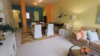 This single-floor space makes a striking first impression with its bold, multi-colored walls in shades of teal, orange, and golden yellow, creating a vibrant and energetic atmosphere throughout. The open layout connects the dining and living areas seamlessly, lending the interior a spacious, airy feel.

The dining area serves as a focal point, anchored by a dark wood dining table surrounded by elegantly slipcovered white chairs. A patterned area rug with warm, earthy botanical motifs defines the dining zone and adds texture underfoot. A dark console table along the wall provides functional surface space and display storage, accented by decorative items including tall dried branches in a vase.

Track lighting along the ceiling provides overhead illumination, while decorative table lamps contribute warm ambient lighting throughout. An ornate gold-framed mirror adorns one wall, adding a classic contrast to the colorful surroundings. A decorative arched window frame serves as a unique wall accent, enhancing the eclectic character of the space.

The living area, partially visible in the foreground, features neutral upholstered seating that balances the vibrant color palette. Overall, the space exudes an eclectic, cheerful ambiance with a mix of bold color and curated décor.