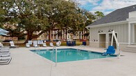 Located at 144 E 62nd St in Savannah, Georgia, this single-story outdoor pool facility offers a well-appointed recreational experience within a residential community setting. The centerpiece of the space is a generously sized swimming pool with clear blue water, accessible via dual entry handrails and equipped with a pool lift for accessibility. The expansive concrete pool deck surrounds the pool on all sides, providing ample space for lounge chairs and outdoor games, including cornhole boards. A striking mature live oak tree anchors the far corner of the property, lending natural shade and a quintessentially Southern ambiance to the outdoor setting. The space is enclosed by a black iron fence for safety and privacy. Adjacent to the pool, a covered porch with ceiling fans and teal-accented doors offers a comfortable shaded retreat with seating. The pool house is clad in board-and-batten siding with gooseneck barn light fixtures and houses gender-designated restrooms, accessible water fountains, and fire safety equipment. The building's clean white exterior and thoughtful detailing reflect a modern farmhouse aesthetic that complements the surrounding community architecture.
