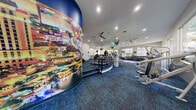 Located at 2121 E Warm Springs Rd in Las Vegas, NV, this well-appointed fitness center offers a generously sized, single-floor exercise space designed to support a comprehensive workout experience. The room is anchored by a striking, curved mural showcasing the iconic Las Vegas Strip, adding a bold and energetic visual identity to the environment. Rich blue patterned carpet covers the floor throughout, complementing the bright white walls and recessed lighting overhead.

The cardio section is well-equipped, featuring multiple treadmills, stair climbers, and other aerobic machines arranged with ample spacing for comfortable use. Ceiling fans are mounted above to promote airflow throughout the space. Multiple flat-screen televisions are suspended from the ceiling, ensuring entertainment is accessible from virtually every area of the room.

The strength training section offers a robust selection of equipment, including cable machines, a Smith machine, plate-loaded resistance machines, and a dedicated free weights area stocked with a full rack of dumbbells and medicine balls. TRX suspension trainers and a heavy bag add functional training options.

Large windows line multiple walls, inviting generous natural light and framing views of the surrounding greenery and palm trees. Full-length mirrors in the strength area further enhance the sense of openness. The overall ambiance is energetic, modern, and thoughtfully equipped for a full-spectrum fitness routine.