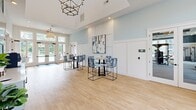 Located at 601 McCaw Bridge Street in Raleigh, NC, this generously sized single-floor community clubhouse offers a thoughtfully designed layout across a broad, open footprint. The entrance greets visitors with soaring ceilings, large windows flooding the space with natural light, light wood flooring, and a statement chandelier, establishing an inviting and polished first impression. The combined dining and living area continues the bright, airy aesthetic, with expansive floor-to-ceiling windows overlooking an outdoor pool and terrace. The living portion features comfortable neutral seating set against a wall of windows, while the dining area includes round wood tables paired with contemporary navy chairs. The kitchen is a standout feature, boasting light gray cabinetry with matte black hardware, stone countertops, and a bold navy chevron-patterned tile backsplash accented by floating natural wood shelves. The hallway connects the various areas seamlessly with clean white wainscoting and warm flooring throughout. The fitness center is expansive and well-appointed, featuring deep navy accent walls, floor-to-ceiling mirrors, recessed lighting, a coffered ceiling detail with wood beam accents, and an extensive array of equipment including free weights, medicine balls, resistance machines, and benches, all set beside large windows with outdoor views.