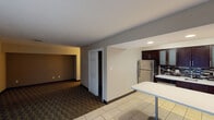 Located at 8751 Michigan Rd in Indianapolis, Indiana, this single-floor suite offers a functional and comfortable layout designed for extended stays or self-sufficient living. The open-concept design connects the kitchen and living areas seamlessly, creating a practical and welcoming flow throughout the space.

The kitchen is a standout feature, outfitted with full-size stainless steel appliances including a refrigerator, dishwasher, and microwave. Rich dark cabinetry with frosted glass accents reaches floor to ceiling, providing generous storage, while a mosaic tile backsplash adds a modern design touch. Light countertops and tile flooring complete the polished look.

The living area transitions between light tile and patterned carpet, giving each zone a distinct character. A sliding glass door fitted with vertical blinds allows natural light to filter in and opens to an outdoor view, offering a bright and airy atmosphere. The space also includes a closet for additional storage.

The bathroom is clean and efficiently designed, featuring a combination tub and shower with a safety grab bar, tile flooring, and a wide vanity with a backlit mirror and integrated hair dryer. The neutral color palette throughout the suite — warm beige walls, white trim, and dark accents — lends a cohesive and understated aesthetic suited for both short and extended occupancy.