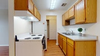 This single-floor residence offers a functional and comfortable layout with consistent design elements throughout. The galley-style kitchen is well-equipped, featuring warm honey-toned wood cabinetry on both sides, white appliances including a coil-top range and refrigerator, light countertops, a stainless steel sink, and a range hood overhead. Fluorescent lighting illuminates the space, and hardwood-style flooring runs underfoot, leading through to a closeted storage area beyond. The living area is open and airy, showcasing the same warm hardwood-style flooring, bright white walls, and a ceiling fan with light fixture. A large window allows natural light to fill the room, offering views of the surrounding neighborhood. Baseboard heating runs along the perimeter, and a wall-mounted air conditioning unit ensures year-round comfort. The bedroom mirrors this clean aesthetic, with hardwood-style flooring, neutral walls, a window with blinds, and baseboard heating. The bathroom is neatly appointed with a wood-vanity, a full tub and shower combo with a striped curtain, and durable tile flooring in an earthy tone. Overall, the space presents a clean, practical, and inviting atmosphere with cohesive finishes throughout.