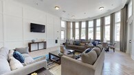 This single-story clubhouse offers a thoughtfully designed communal lounge that blends comfort and sophistication. The space is anchored by a striking curved wall of large bay windows dressed with floor-length patterned drapes, flooding the interior with abundant natural light and offering views of the surrounding outdoors. Crown molding and white wainscoting panels line the walls, lending a refined architectural character to the room.

The lounge is furnished with plush, neutral-toned sofas and accent chairs arranged around a dark wood coffee table, creating an inviting gathering area. Colorful area rugs in blue, gold, and gray tones add visual interest and warmth to the light hardwood-style flooring beneath. A wall-mounted flat-screen television is integrated into a paneled accent wall, providing a dedicated entertainment focal point complemented by a slim console table beneath.

Recessed and flush-mount ceiling fixtures provide consistent ambient lighting throughout, ensuring the space remains bright and welcoming at any time of day. The overall ambiance is polished yet comfortable, making the clubhouse an ideal setting for socializing and relaxation.