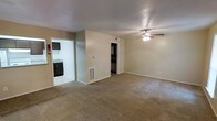 This single-floor residence offers a straightforward and functional layout designed for comfortable everyday living. The main living area is generously sized, featuring wall-to-wall carpeting in a neutral beige tone that adds warmth underfoot. A ceiling fan with integrated lighting is centrally mounted overhead, providing both air circulation and ambient light to the space. The walls are finished in a soft, light beige palette that creates a clean, understated backdrop throughout.

Adjacent to the living area, a tiled kitchen is visible through a wide doorway, outfitted with dark cabinetry and dark appliances that offer a contrasting accent to the otherwise light interior. A floor vent and a wall-mounted thermostat are thoughtfully placed for climate control convenience.

Toward the back of the living space, a second doorway opens into what appears to be a walk-in closet or storage area, also featuring dark cabinetry or shelving, adding practical storage capacity to the unit. The textured ceiling runs consistently throughout, and the clean white trim framing each doorway provides a crisp, finished look. Overall, this space presents a tidy and functional environment with a neutral, move-in-ready aesthetic suitable for a variety of living arrangements.