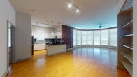 This single-floor residence showcases a thoughtful blend of industrial character and modern comfort. The living area is defined by a dramatic curved wall of floor-to-ceiling windows fitted with blinds, flooding the space with natural light and creating an airy, open atmosphere. Warm hardwood flooring extends throughout the main living and kitchen areas, complemented by a striking exposed brick accent wall that adds texture and character. A ceiling fan anchors the curved living space, enhancing both comfort and style.

The open kitchen is well-appointed with white cabinetry, dark granite countertops, a tile backsplash, and stainless steel appliances including a refrigerator and microwave. Pendant lights hang above the kitchen island, which also serves as a natural divider between the cooking and living areas. Track lighting runs across the kitchen ceiling for functional illumination.

The bedroom offers plush carpet flooring, dual windows for natural light, and a ceiling fan overhead. The bathroom includes a full tub-shower combination with white tile surround, a single vanity with storage, and tile flooring. A generously sized walk-in closet with wire shelving provides ample storage. A dedicated laundry area houses a full-size top-loading washer and dryer with additional shelving above.