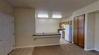 This single-floor space is designed with practicality in mind, centering around an open-concept kitchen that connects seamlessly to the living area. The kitchen is anchored by a peninsula counter with a dark laminate surface and an integrated sink, functioning as both a workspace and a casual dining or serving bar. Warm honey-toned wood cabinetry lines the kitchen walls, providing ample storage above and below the countertops. Standard white appliances, including a gas range with overhead ventilation and a full-sized refrigerator, are neatly arranged along the back wall, ensuring a fully equipped cooking space.

Overhead fluorescent lighting illuminates the kitchen area efficiently, keeping the space well-lit for everyday tasks. The flooring transitions from tile in the kitchen and entry zone to carpet in the adjacent living area, visually separating the two functional spaces while maintaining an open feel. The neutral beige walls and light-colored ceiling contribute to a clean, understated aesthetic throughout. A full-height door on the far side of the kitchen suggests access to additional areas of the unit, adding to the overall sense of a connected, single-level floor plan.