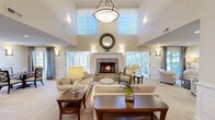 Located on Wildwood Drive in Menomonee Falls, Wisconsin, this generously sized single-story community facility offers a refined and welcoming atmosphere across its expansive floor plan. The centerpiece of the space is a dramatic great room featuring soaring two-story ceilings, clerestory windows that flood the interior with natural light, and a tile-surround fireplace framed by an elegant mantle. Comfortable seating arrangements and wainscoting details lend the room a warm, sophisticated ambiance. Adjacent to the lounge is a formal dining nook with wainscoted walls, framed artwork, and garden-facing windows. The open-concept kitchen is equipped with rich cherry cabinetry, granite countertops, stone tile backsplash, and stainless steel appliances, connected seamlessly to a casual dining area. A dedicated ballroom showcases stunning inlaid parquet hardwood flooring, a vaulted tray ceiling, and a mirrored disco ball, making it ideal for events and entertainment. The multi-purpose event room features large windows on three sides, carpeted flooring, and ample space for banquet-style seating. Throughout, wide corridors with decorative built-ins, nature-inspired wall art, and abundant glazing create bright, elegant transitions between spaces. The overall design balances classic architectural details with functional, community-focused versatility.