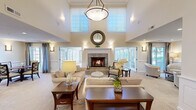 Located on Wildwood Drive in Menomonee Falls, Wisconsin, this generously sized single-story community facility offers a refined and welcoming atmosphere across its expansive floor plan. The centerpiece of the space is a dramatic great room featuring soaring two-story ceilings, clerestory windows that flood the interior with natural light, and a tile-surround fireplace framed by an elegant mantle. Comfortable seating arrangements and wainscoting details lend the room a warm, sophisticated ambiance. Adjacent to the lounge is a formal dining nook with wainscoted walls, framed artwork, and garden-facing windows. The open-concept kitchen is equipped with rich cherry cabinetry, granite countertops, stone tile backsplash, and stainless steel appliances, connected seamlessly to a casual dining area. A dedicated ballroom showcases stunning inlaid parquet hardwood flooring, a vaulted tray ceiling, and a mirrored disco ball, making it ideal for events and entertainment. The multi-purpose event room features large windows on three sides, carpeted flooring, and ample space for banquet-style seating. Throughout, wide corridors with decorative built-ins, nature-inspired wall art, and abundant glazing create bright, elegant transitions between spaces. The overall design balances classic architectural details with functional, community-focused versatility.