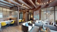 This single-story space makes a striking impression through its masterful use of natural wood throughout. Exposed ceiling beams run the length of the interior, complemented by rich wood-paneled walls and wide-plank hardwood flooring that create a cohesive, warm aesthetic. Structural columns anchor the open layout, adding architectural character while defining distinct zones within the space.

The lounge area is furnished with dark-framed white upholstered chairs and a neutral sofa, accented by teal and green throw pillows. A large abstract wall piece in muted copper and teal tones serves as a focal point, adding an artistic dimension to the space. An industrial-style chandelier with exposed bulbs provides ambient overhead lighting.

Adjacent to the lounge, a dining or collaborative area features vibrant yellow chairs paired with sleek tables, offering a bold contrast to the warm wood tones. Steel-framed glass partitions separate this area while maintaining visual openness. A private room with a glass door is accessible beyond, providing an enclosed retreat within the larger layout. Together, these elements create a sophisticated, inviting atmosphere with a distinctly upscale lodge-inspired character.