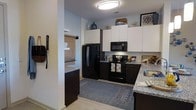 This single-floor residence is designed with a clean, contemporary aesthetic that balances functionality and style. The kitchen serves as a standout feature, showcasing a striking two-tone design with dark espresso lower cabinetry contrasted by crisp white upper cabinets that extend to the ceiling, providing generous storage throughout. Granite countertops with subtle veining add a touch of elegance to the space, while the full suite of black appliances — including a side-by-side refrigerator, over-the-range microwave, and electric range — creates a cohesive, modern look. A flush-mount ceiling light provides bright, even illumination across the kitchen area. Light-toned flooring runs throughout, lending an open and airy feel to the interior. A patterned area rug anchors the kitchen space, adding warmth and visual interest. An open sightline connects the kitchen to an adjacent room, enhancing the sense of flow and spaciousness throughout the floor plan. The overall ambiance is modern and inviting, with thoughtful design details that make this a practical and attractive living environment.