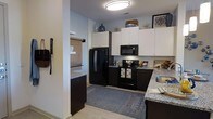 This single-floor residence is designed with a clean, contemporary aesthetic that balances functionality and style. The kitchen serves as a standout feature, showcasing a striking two-tone design with dark espresso lower cabinetry contrasted by crisp white upper cabinets that extend to the ceiling, providing generous storage throughout. Granite countertops with subtle veining add a touch of elegance to the space, while the full suite of black appliances — including a side-by-side refrigerator, over-the-range microwave, and electric range — creates a cohesive, modern look. A flush-mount ceiling light provides bright, even illumination across the kitchen area. Light-toned flooring runs throughout, lending an open and airy feel to the interior. A patterned area rug anchors the kitchen space, adding warmth and visual interest. An open sightline connects the kitchen to an adjacent room, enhancing the sense of flow and spaciousness throughout the floor plan. The overall ambiance is modern and inviting, with thoughtful design details that make this a practical and attractive living environment.