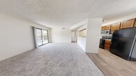 Located at 1001 Tramway Blvd NE in Albuquerque, New Mexico, this single-floor, three-bedroom, one-bathroom unit offers a practical and well-organized layout. The open living room is finished with plush carpet and features a sliding glass door that leads to a balcony, filling the space with natural light and providing outdoor views of the surrounding area. A textured ceiling is consistent throughout the unit, adding a classic residential character.

The kitchen is efficiently designed with warm wood-toned cabinetry, wood-look vinyl flooring, black appliances including a refrigerator and electric range, a stainless steel microwave, and a pendant light fixture overhead. A half-wall pass-through connects the kitchen to the living area, creating an open, communicative flow between the two spaces.

The three bedrooms are carpeted and bright, each with white walls and textured ceilings. The primary bedroom offers a spacious feel with a sliding closet for storage and a window that brings in natural light. The additional bedrooms are similarly finished and versatile in use.

The bathroom is clean and functional, featuring wood-look vinyl flooring, a single-sink vanity with a warm wood cabinet, a full tub-shower combination with brushed nickel fixtures, and a recessed medicine cabinet.