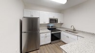 This single-floor unit offers a clean, modern aesthetic with thoughtfully updated finishes throughout. The kitchen is a standout feature, showcasing white shaker-style cabinetry, speckled granite countertops, and a full suite of stainless steel appliances including a refrigerator, electric range, over-the-range microwave, and dishwasher. A stainless steel undermount sink with a brushed nickel faucet completes the functional L-shaped layout, all set atop durable wood-style plank flooring that extends seamlessly into the living areas.

The living and bedroom spaces are bright and open, finished with the same warm-toned hardwood-style flooring and neutral white walls that create a clean, versatile backdrop. Ceiling fans with integrated lighting fixtures are present in multiple rooms, enhancing both comfort and functionality. The bedroom includes a window with blinds allowing for natural light, as well as a closet door providing convenient storage.

The bathroom is well-appointed with a granite-topped vanity, a full soaking tub, and a tiled tub surround featuring large-format stone-look tiles in earthy gray tones that lend a spa-like quality to the space. Brushed nickel fixtures and a towel ring add cohesive, contemporary detailing. Overall, the unit presents a fresh and updated living environment with consistent, modern finishes.