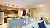 This well-appointed single-floor residence offers a thoughtfully designed open-concept layout that connects the living and kitchen areas in a cohesive, flowing arrangement. The kitchen is a standout feature, showcasing dark cabinetry with a rich wood finish, granite countertops, and a peninsula that doubles as a breakfast bar — ideal for casual dining or entertaining. A decorative wine rack mounted on the wall adds a charming bistro-inspired character, complemented by track lighting overhead that provides warm, focused illumination throughout the space. Built-in shelving offers both functional storage and display opportunities. The living area transitions seamlessly from the kitchen, anchored by a plush white sofa and a mirrored coffee table that reflects light and adds a sense of openness. Decorative glass vases with vibrant floral arrangements bring a lively pop of color to the space. The neutral, earthy wall tones throughout create a calm and inviting ambiance, while the overall design balances modern aesthetics with warm, personalized touches. The single-level layout ensures easy accessibility and a comfortable flow between spaces, making it well-suited for both everyday living and hosting guests.