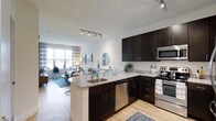 This single-floor residence showcases a thoughtfully designed open-concept layout that seamlessly connects the kitchen and living areas. The kitchen is a standout feature, boasting rich dark cabinetry that contrasts beautifully with light granite countertops, offering ample storage and prep space. A peninsula with an integrated sink extends into the living area, creating a natural gathering point and a clear sightline throughout the space. Stainless steel appliances, including a range, microwave, and dishwasher, complete the well-appointed kitchen. Warm-toned hardwood-style flooring flows continuously throughout, adding a sense of cohesion and warmth. Track lighting along the ceiling provides both ambient and task illumination, complementing the generous natural light that pours in through large windows in the living area. The living space is open and airy, with neutral white walls that allow for flexible décor. Patterned curtains and decorative accents add a touch of personality, while the overall design maintains a clean, contemporary aesthetic. The combination of functional design and modern finishes makes this a welcoming and highly livable space.