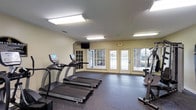 This single-floor fitness center is designed to provide a functional and comfortable workout environment. The space is outfitted with a selection of cardio equipment, including multiple treadmills by TRUE Fitness, as well as a multi-station strength training machine, making it suitable for a well-rounded exercise routine. The flooring consists of durable rubber gym matting, ideal for both cardio and strength training activities. Overhead fluorescent lighting ensures the room is brightly and evenly lit throughout. The room's walls are finished in a neutral cream tone, contributing to a clean and open atmosphere. A wall-mounted television is positioned for easy viewing during workouts. Notably, the space features several large, white-framed glass-paneled doors and windows along one wall, allowing natural light to fill the room while providing views of and direct access to an outdoor area. An illuminated exit sign and proper ventilation are also present, reflecting attention to safety and comfort. Overall, the fitness center offers a practical, well-maintained space that balances functionality with a welcoming, light-filled ambiance.