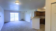 This single-floor residence offers a practical and well-organized open-concept layout that seamlessly connects the living area and kitchen. The living room is spacious and comfortable, featuring wall-to-wall carpeting that adds warmth underfoot, a large sliding glass door with vertical blinds that floods the space with natural light, and a floor vent heating system for year-round comfort. A doorway visible from the main living area provides access to additional rooms, hinting at a thoughtful floor plan designed for ease of movement. The kitchen is clearly defined by a peninsula-style countertop that serves as a natural divider between the cooking and living spaces. Rich, warm-toned wooden cabinetry lines the upper walls, offering generous storage, while the flooring transitions to tile in the kitchen area, providing a practical and easy-to-clean surface. A ceiling-mounted flush light fixture illuminates the shared space, and ventilation is supported by ceiling-integrated HVAC vents throughout. The overall ambiance is clean, neutral, and move-in ready, with white walls and a straightforward layout that offers a welcoming, functional living environment.